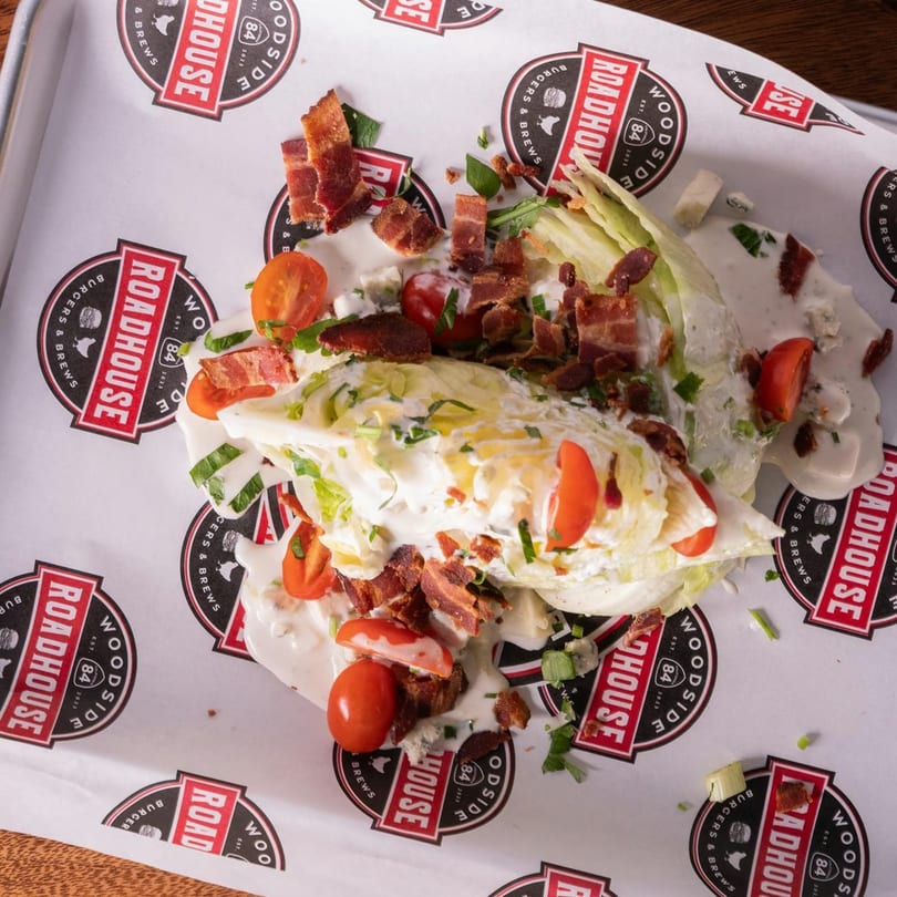 Woodside Roadhouse wedge salad with bacon, tomatoes, and blue cheese dressing on branded paper.