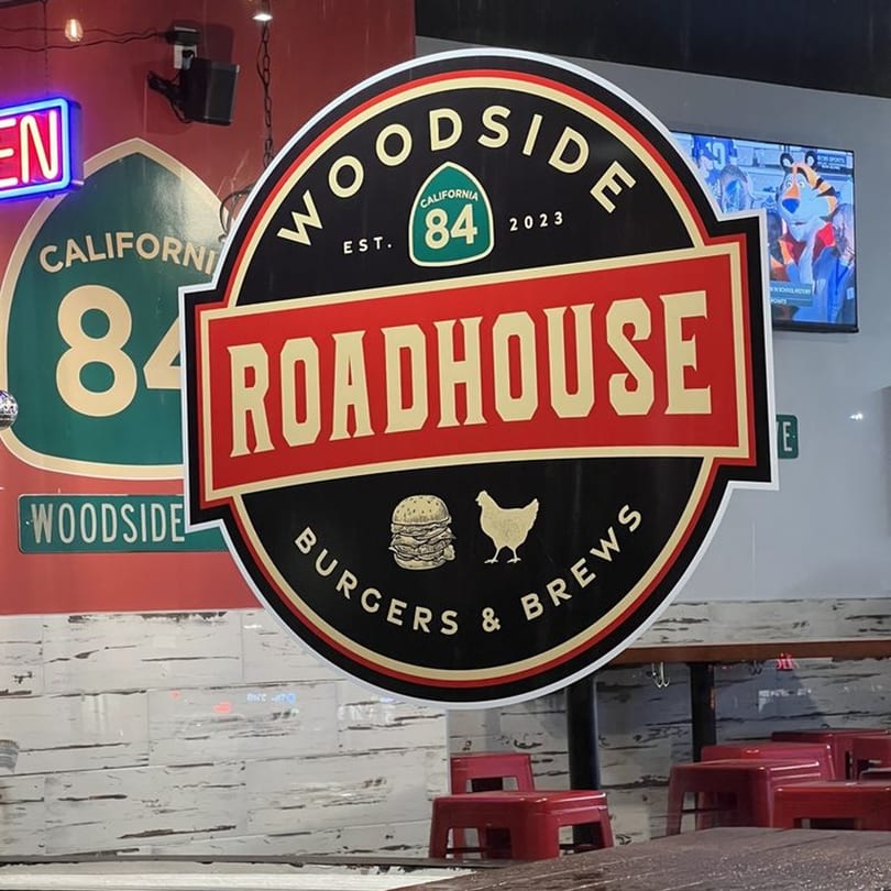 Woodside Roadhouse sign with a burger and chicken logo, visible through a window with red stools.